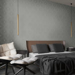 A modern bedroom features a large bed with a brown leather headboard and dark pillows. Two sleek pendant lights illuminate a cushioned chair against metallic York Wallcoverings' Light Grey & Silver Harlowe Wallpaper, enhancing the beige wall's subtle diamond pattern.