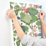 A person is hanging a sheet of York Wallcoverings' Lotus Pond Sky/ Multi Wallpaper Purple, Pink (60 Sq.Ft.) with a botanical pattern featuring large green leaves and pink flowers. The individual's hands are visible, adjusting the high-performance substrate on a white wall. The person wears a striped long-sleeve shirt and a ring on one finger.