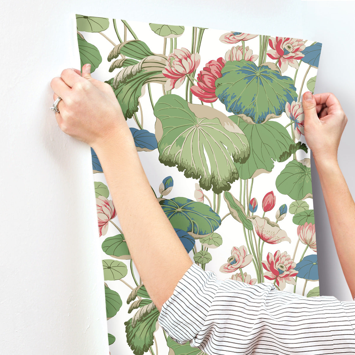 A person is hanging a sheet of York Wallcoverings' Lotus Pond Sky/ Multi Wallpaper Purple, Pink (60 Sq.Ft.) with a botanical pattern featuring large green leaves and pink flowers. The individual's hands are visible, adjusting the high-performance substrate on a white wall. The person wears a striped long-sleeve shirt and a ring on one finger.