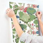 A person, dressed in a long-sleeved striped shirt and a ring on their left hand, is applying York Wallcoverings Lotus Pond Heather/Cotton Wallpaper Grey, Blue (60 Sq.Ft.) with a floral and leaf pattern. The green leaves and pink flowers create a tranquil design against the white wall, showcasing the ease of installation.