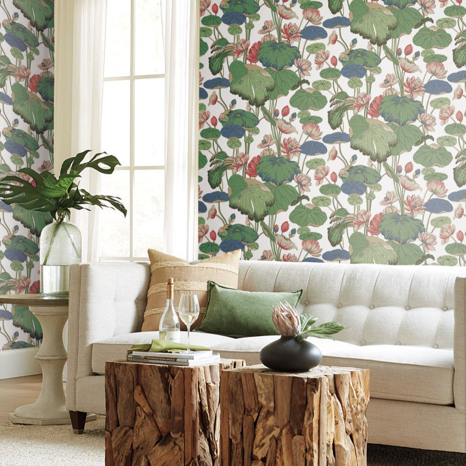 A cozy living room with a light gray tufted sofa adorned with green and beige throw pillows. The space features York Wallcoverings' Lotus Pond Sky/ Multi Wallpaper Purple, Pink (60 Sq.Ft.), showcasing large green leaves and red flowers for easy installation. A rustic wooden coffee table holds a glass, a plant, and a small vase.