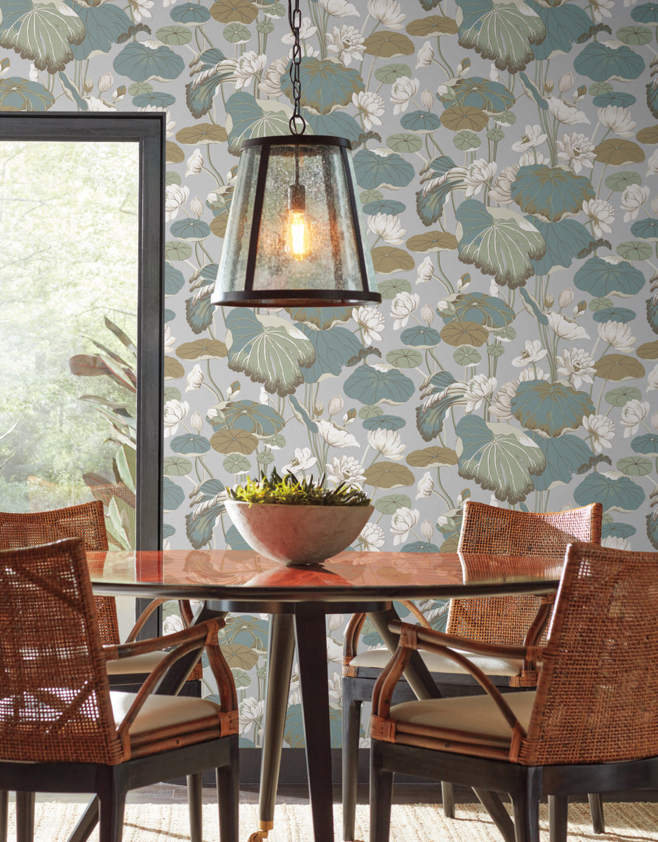 A dining area with a circular wooden table, four wicker chairs, and a metal-framed pendant light. There's a decorative bowl with greenery on the table. The wall features Lotu Pond Heather/Cotton Wallpaper Grey, Blue (60 Sq.Ft.) by York Wallcoverings in blue, green, and white hues, offering a tranquil design through the large window's outside view.