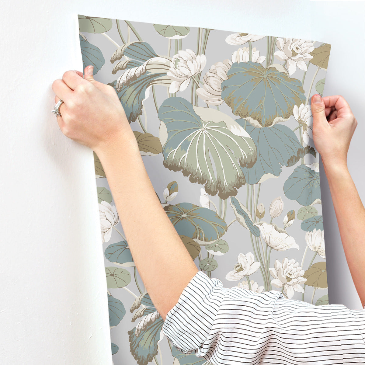 A person in a striped shirt is attaching a piece of Lotus Pond Heather/Cotton Wallpaper Grey, Blue (60 Sq.Ft.) by York Wallcoverings featuring large, tranquil botanical prints with muted colors, including shades of green, blue, and beige, to a white wall. Their hands are visible, holding the wallpaper by the corners for easy installation.