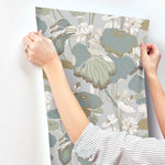 A person in a striped shirt is attaching a piece of Lotus Pond Heather/Cotton Wallpaper Grey, Blue (60 Sq.Ft.) by York Wallcoverings featuring large, tranquil botanical prints with muted colors, including shades of green, blue, and beige, to a white wall. Their hands are visible, holding the wallpaper by the corners for easy installation.