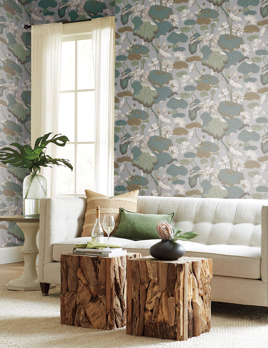 A cozy living room with a light gray tufted sofa, green and beige pillows, and two rustic wooden coffee tables. Behind the sofa is a large window with white curtains. The room features York Wallcoverings Lotus Pond Cotton/Peacock Wallpaper Green, Pink (60 Sq.Ft.) with a floral pattern in green, blue, and neutral tones for easy installation and removal.