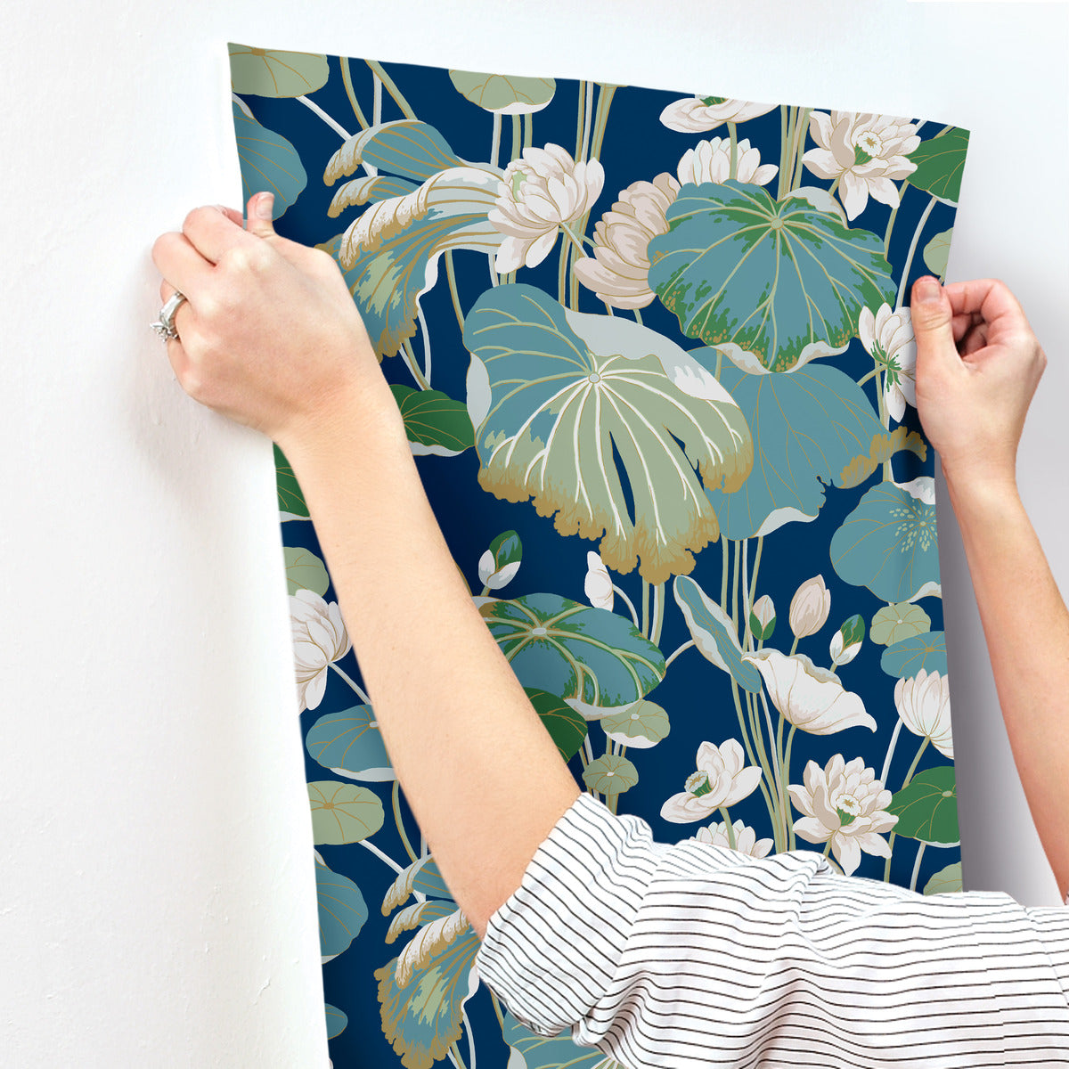 A person wearing a striped shirt is hanging York Wallcoverings Lotus Pond Sky/ Multi Wallpaper Purple, Pink (60 Sq.Ft.) with a botanical print featuring large green leaves and white flowers on a dark blue background. The person’s hands are visible as they smooth out the paper, made from high performance substrate, against a white wall.