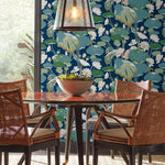 A cozy dining area with a round wooden table and four wicker chairs is set against tranquil Lotus Pond Heather/Cotton Wallpaper Grey, Blue (60 Sq.Ft.) by York Wallcoverings, featuring lotus leaves and blossoms. A modern pendant light hangs above the table, illuminating a decorative bowl filled with greenery. Large windows provide natural light.