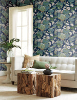 A cozy living room features a beige tufted sofa with green and white cushions, wooden side tables, and a large green plant in a vase. The backdrop wall is adorned with Lotus Pond Heather/Cotton Wallpaper Grey, Blue (60 Sq.Ft.) by York Wallcoverings in shades of blue, green, and white, complemented by a tall window with sheer curtains.