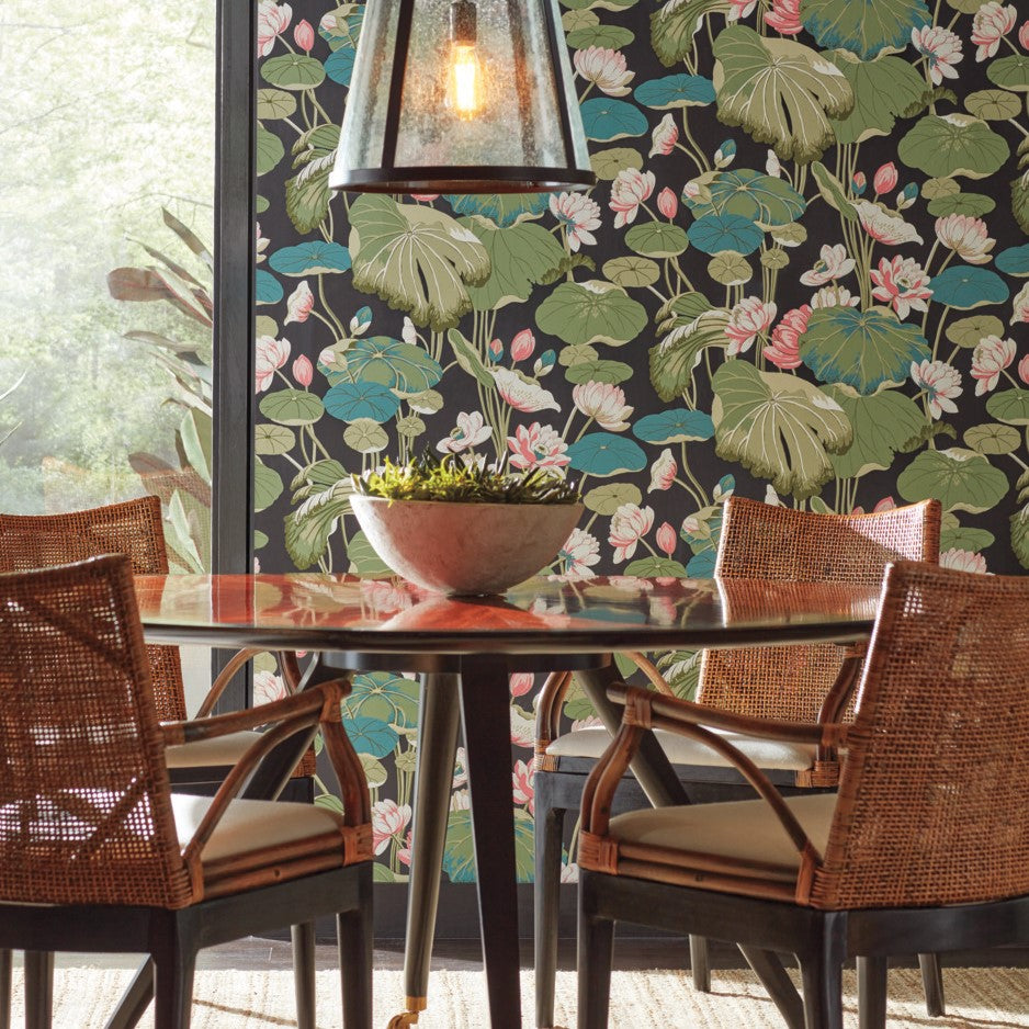 A dining area features a round wooden table with four wicker chairs. A large potted plant rests on the table, while the room's backdrop is a wall adorned with vibrant Lotus Pond Sky/ Multi Wallpaper Purple, Pink (60 Sq.Ft.) by York Wallcoverings. An industrial-style pendant light hangs overhead, highlighting the easy installation and high-performance substrate of the decor.