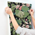 A person with a striped shirt hangs a sheet of York Wallcoverings' Lotus Pond Sky/ Multi Wallpaper Purple, Pink (60 Sq.Ft.) featuring large green leaves and pink flowers on a white wall. The wallpaper has a black background, and the person's hands are visible holding it in place. Enjoy easy installation thanks to its high performance substrate.