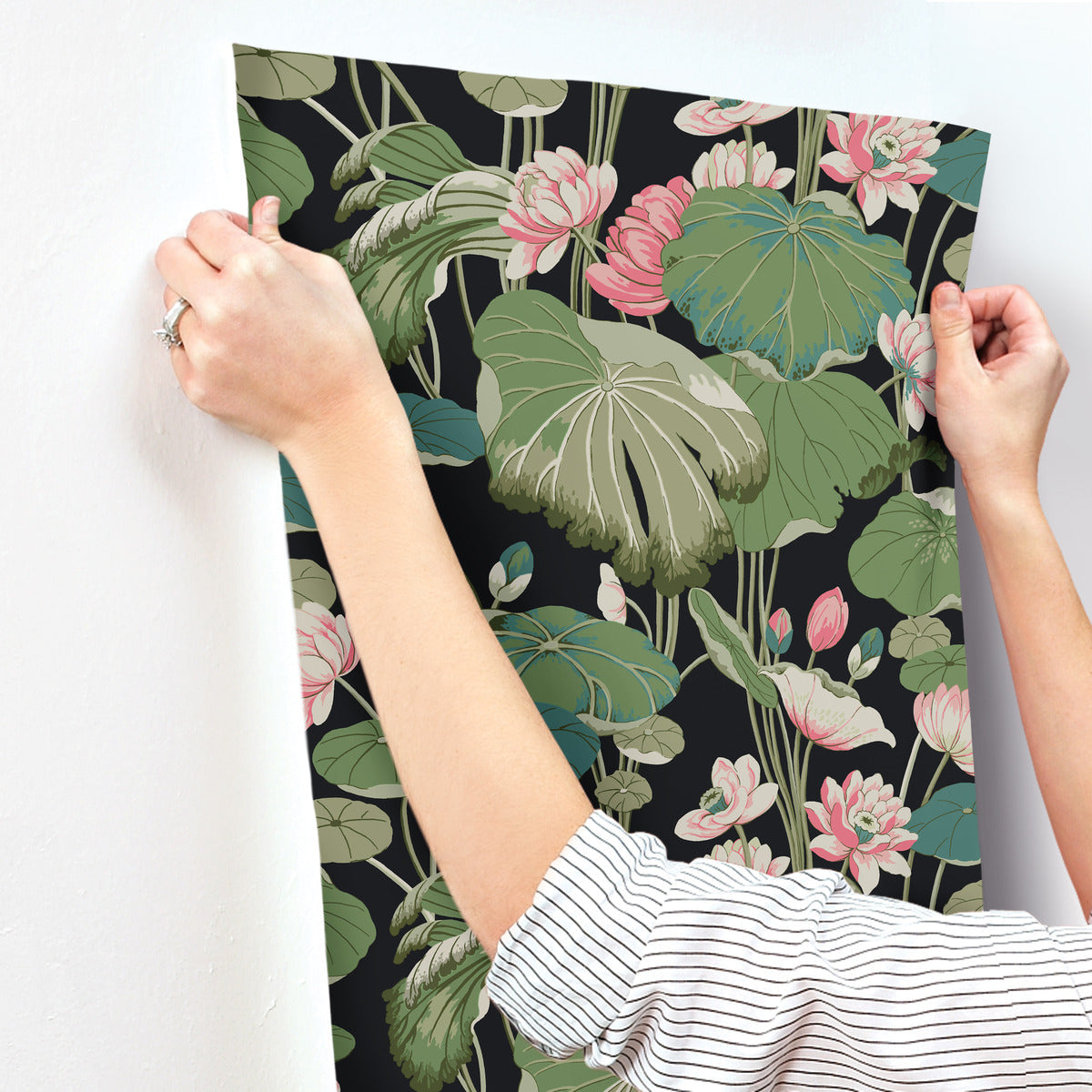 A person with a striped shirt and a ring on their left hand is installing York Wallcoverings Lotus Pond Cotton/Peacock Wallpaper Green, Pink (60 Sq.Ft.). This floral wallpaper with a dark background features large green leaves and pink flowers with rounded petals, creating a lush and vibrant design, making for easy installation and removal.