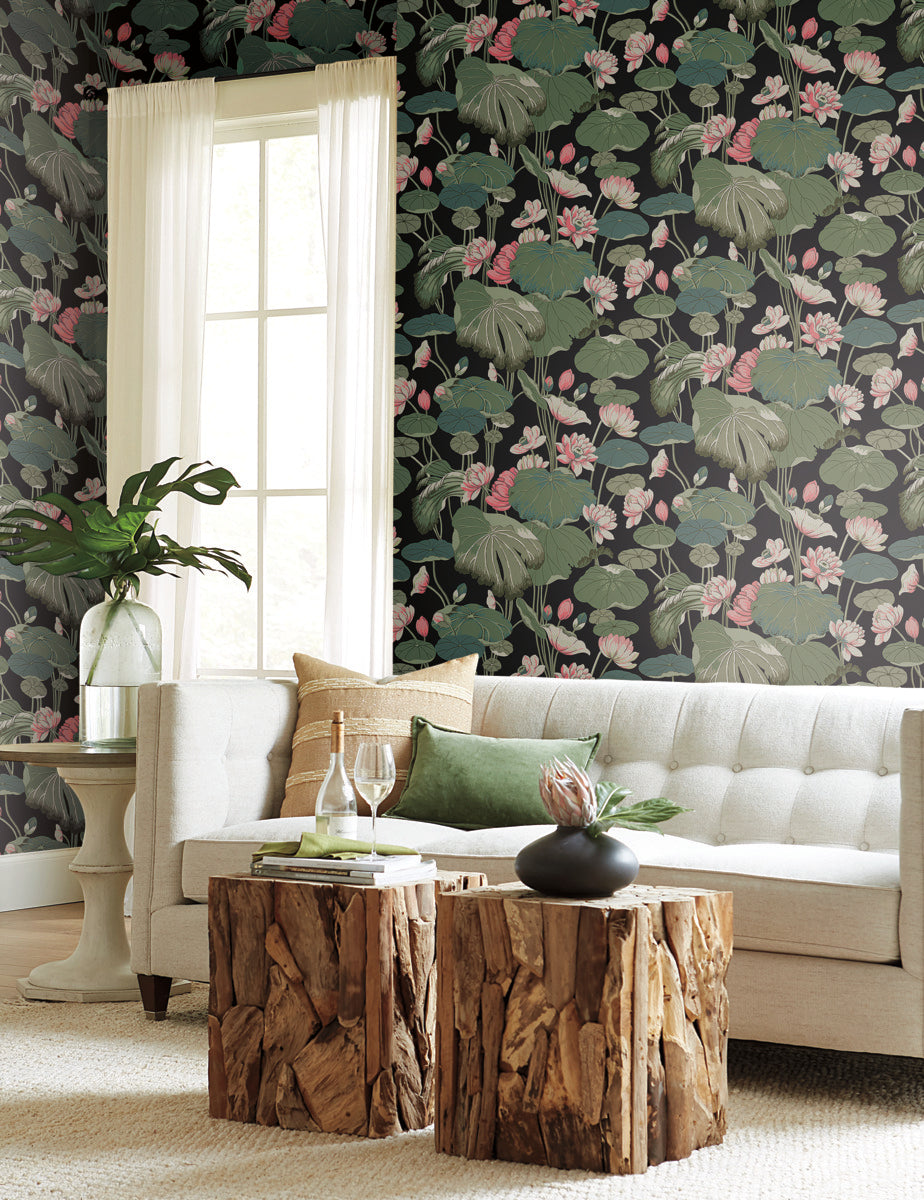 A cozy living room with a cream-colored sofa, green and beige throw pillows, and two rustic wooden coffee tables. The wall behind features York Wallcoverings Lotus Pond Cotton/Peacock Wallpaper Green, Pink (60 Sq.Ft.) for easy installation and removal, while a tall window with sheer curtains allows natural light in. A plant and decorative items adorn the space.