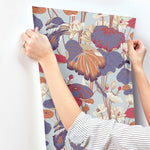 A person is holding up a piece of Lotus Pond Sky/ Multi Wallpaper Purple, Pink (60 Sq.Ft.) by York Wallcoverings against a white wall. The wallpaper, known for its easy installation, features large, colorful leaves and flowers in shades of orange, purple, and beige on a light blue background. The person's sleeves are white with black stripes.