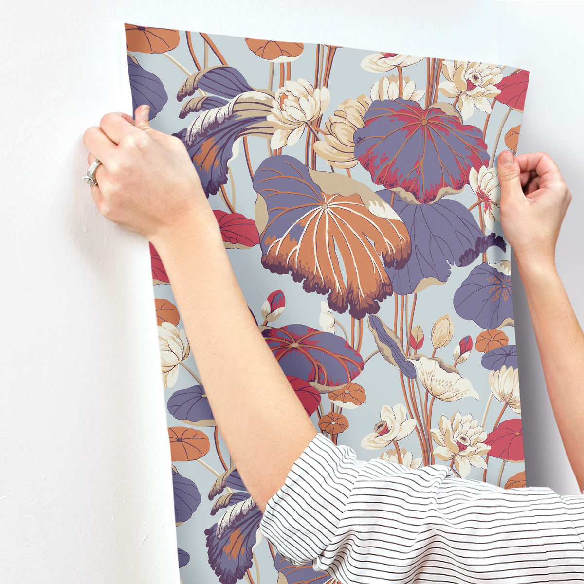 A person is holding and aligning a Lotus Pond Cotton/Peacock Wallpaper Green, Pink (60 Sq.Ft.) by York Wallcoverings with a botanical and floral design on a wall. The wallpaper features large, colorful flowers and leaves in shades of orange, purple, and beige. The person is wearing a striped long-sleeve shirt and a ring on one hand.