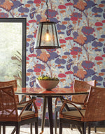 A modern dining area with a round wooden table and four wicker chairs. A metal pendant lamp hangs above the table, and a bowl of plants serves as a centerpiece. The wall features tranquil design elements with York Wallcoverings' Lotus Pond Heather/Cotton Wallpaper Grey, Blue (60 Sq.Ft.) in colorful floral hues of red, purple, and cream.