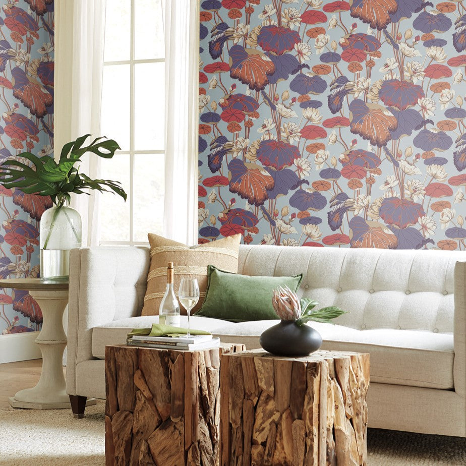 A cozy living room with a beige sofa, green pillows, and a wooden stump coffee table. The space features York Wallcoverings Lotus Pond Sky/Multi Wallpaper Purple, Pink (60 Sq.Ft.) in vibrant red, blue, and orange hues. A large glass vase with greenery sits beside the sofa near a window with white curtains.