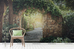 A serene garden scene with a stone archway leading to a Decor2Go Wallpaper Mural path, framed by lush green foliage. A wicker chair with a green cushion sits in the foreground.