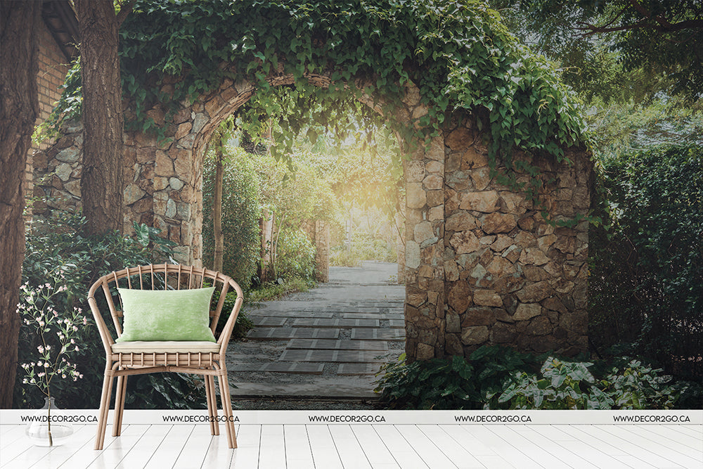A serene garden scene with a stone archway leading to a Decor2Go Wallpaper Mural path, framed by lush green foliage. A wicker chair with a green cushion sits in the foreground.