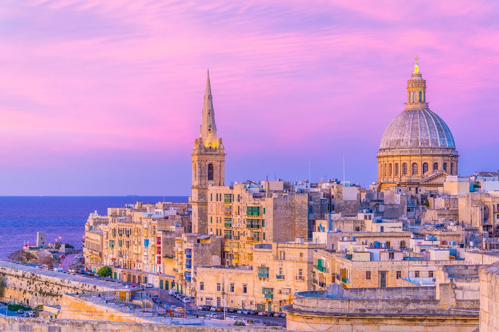 A picturesque view of Valletta, Malta at sunset, featuring historic architectural design with prominent church domes and spires against a pink and purple sky can be captured perfectly with the Malta View Wallpaper Mural by Decor2Go Wallpaper Mural.