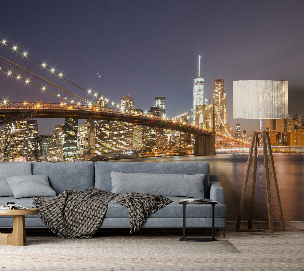A contemporary living room with a large gray sofa and floor lamp, superimposed against a panoramic city view mural of the Brooklyn Bridge and Manhattan skyline featuring the Decor2Go Wallpaper Mural's Manhattan's Night Sky Wallpaper Mural.
