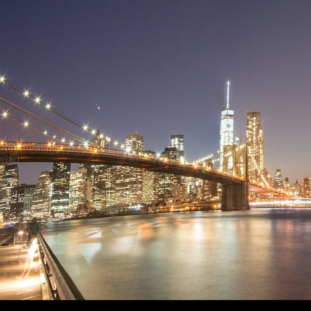 Panoramic view of New York City at night showcasing Brooklyn bridges to the left and Manhattan Bridge to the right with illuminated skyscrapers and reflected city lights in the water using Decor2Go Wallpaper Mural's Manhattan's Night Sky Wallpaper Mural.