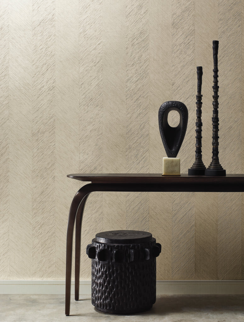 A minimalist interior featuring a sleek, dark wooden table with an African-inspired sculpture, tall black metal candlesticks using Decor2Go Winnipeg's Metallic Chevron Wallpaper Cream as the backdrop, and a textured black stool against a subtly patterned.