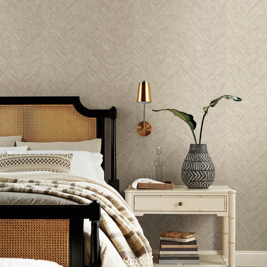 The bedroom interior features a wicker and wood bed frame, patterned pillows, and a beige blanket. A white nightstand displays a geometric vase with green leaves, books, and a towel. Mounted above the nightstand is a brass wall light. The room's texture and modern flair are enhanced by the Cream Metallic Chevron Wallpaper from Decor2Go Wallpaper Mural.