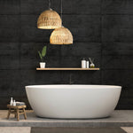 A modern bathroom showcasing a white oval bathtub against the Midnight Slab Wallpaper Mural by Decor2Go exudes urban aesthetic flair. Two wicker pendant lights hang above, while a wooden shelf holds a potted plant and toiletries. A small wooden stool adorned with decorative items sits beside the tub, enhancing the 3D realism of the space.