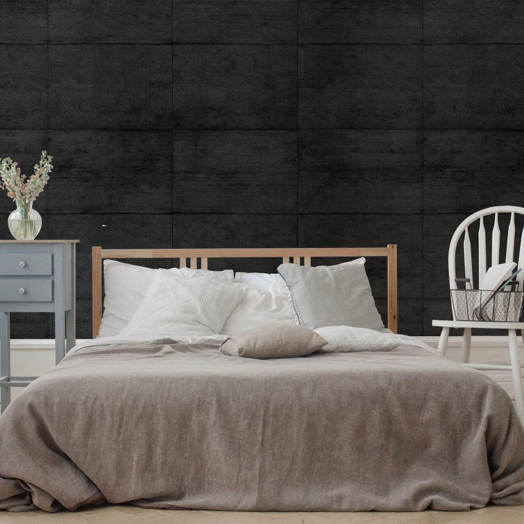 A minimalist bedroom features a wooden bed frame with white and beige bedding against the Midnight Slab Wallpaper Mural by Decor2Go, which provides a dark concrete-textured wall. A small blue side table adorned with flowers and a white chair paired with a basket are positioned beside the bed, adding to the urban aesthetic.