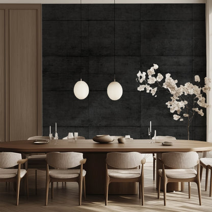 A modern dining room with a long wooden table surrounded by beige chairs exudes an urban aesthetic. Two spherical pendant lights hang above the table, casting a warm glow against the dark, Midnight Slab Wallpaper Mural by Decor2Go, while decorative dried branches in a vase complete the minimalist look.