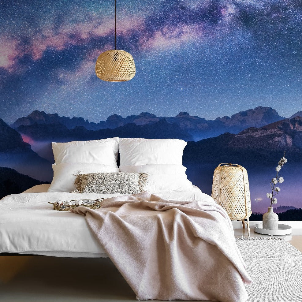 A luxurious bedroom with a large white bed draped with a beige blanket. The room features a stunning Milky Way Mountains Wallpaper Mural from Decor2Go Wallpaper Mural. Wicker lamps add a cozy ambiance.