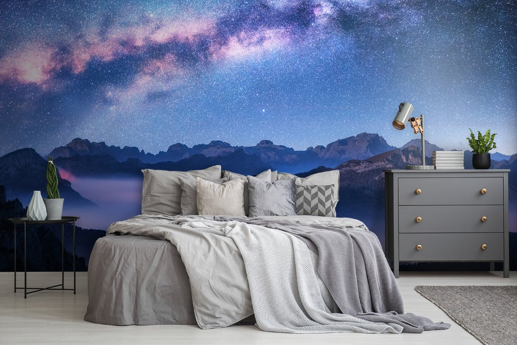 A cozy bedroom featuring a bed with gray linens under a vibrant Decor2Go Wallpaper Mural with mountain landscape. A side table, lamp, gray dresser, and decorative plants complement the serene setting.