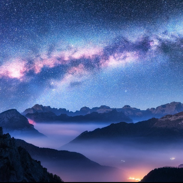 A breathtaking night sky filled with stars over misty, mountainous terrain, featuring the Milky Way Mountains Wallpaper Mural and lights from a distant town below by Decor2Go Wallpaper Mural.