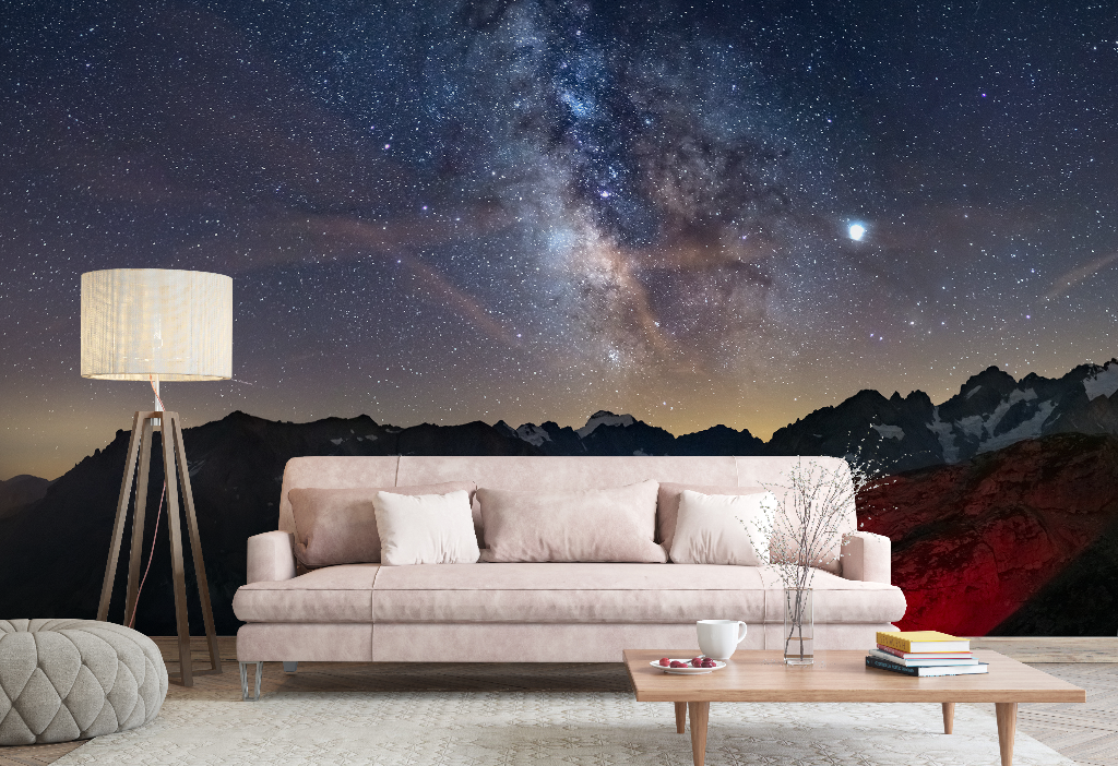 A stylish living room with a beige sofa, wooden coffee table, and floor lamp, set against a large Decor2Go Wallpaper Mural of the Milky Way and mountainous landscape.