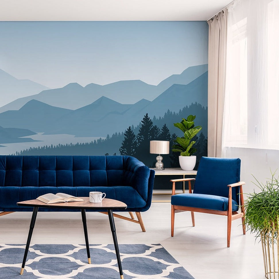 A stylish living room with a large feature wall of Misty Blue Mountains Wallpaper Mural from Decor2Go Wallpaper Mural and a lake, furnished with a navy blue sofa, matching armchair, and a dark wood coffee table with open books.