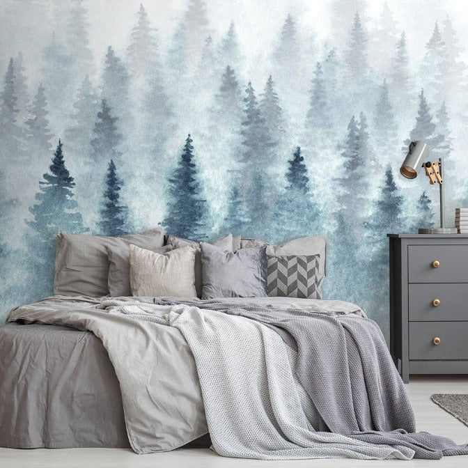 A cozy bedroom featuring a double bed with gray bedding, situated against a Decor2Go Wallpaper Mural of Misty Tree Tops. A rustic wood dresser and black side table with a plant and lamp complete the scene.