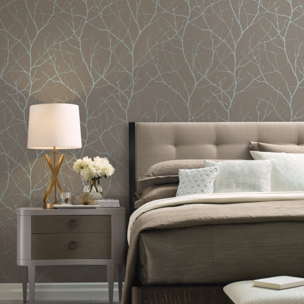 A stylish bedroom features a gray upholstered bed with beige bedding and decorative pillows. A modern lamp and bouquet sit on the nightstand, enhanced by York Wallcoverings' Mocha & Silver Trees Silhouette Wallpaper, showcasing an elegant light-colored tree branch pattern.