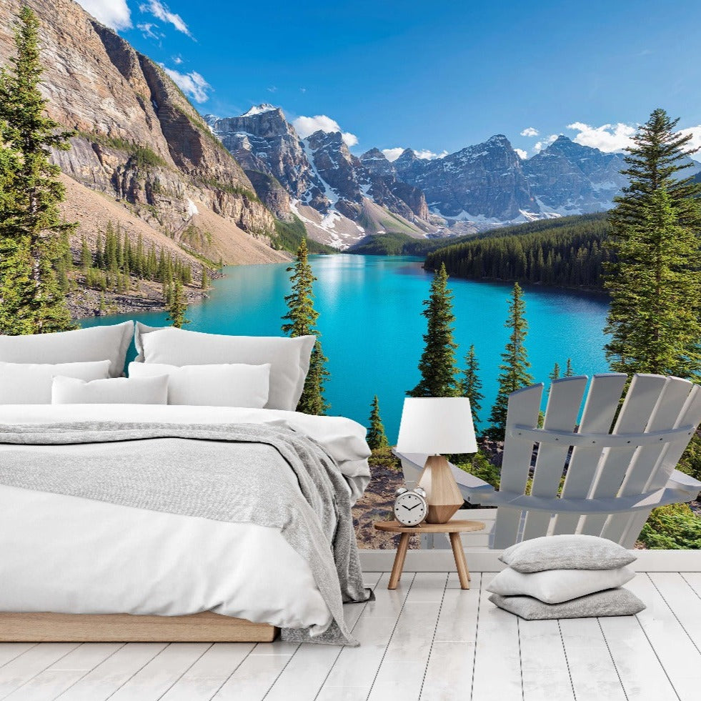 A modern bedroom with an open wall featuring a Decor2Go Wallpaper Mural of Moraine Lake surrounded by forested mountains. The room includes a neat bed, side table, chair, and a