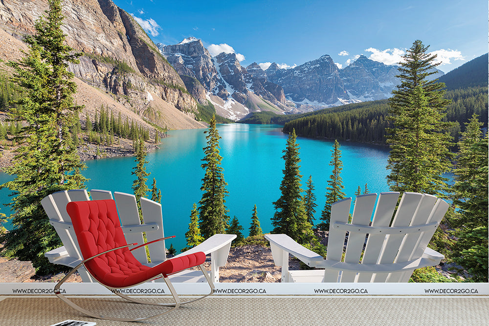 Two Adirondack chairs facing the vivid imagery of Moraine Lake Wallpaper Mural, surrounded by lush green pine trees and majestic mountains in a clear sunny setting.