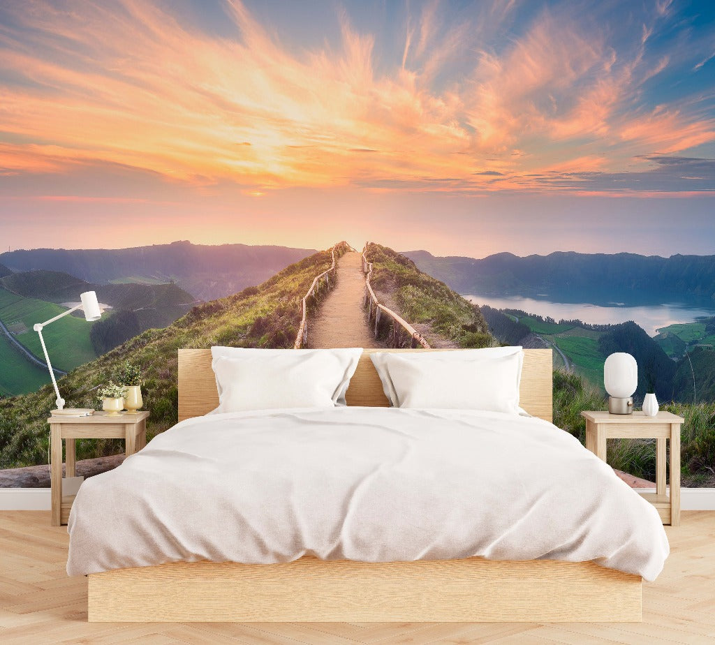 A serene bedroom with a large bed facing a wall-sized Decor2Go Wallpaper Mural of a Mountaintop Trail overlooking a mountain pathway leading into a sunset with distant water views.