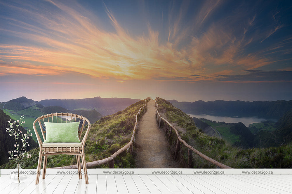 A serene room blending with nature; a wicker chair with a green cushion beside a small plant, in front of a Decor2Go Wallpaper Mural of a sunset over a Mountaintop Trail and a lake.