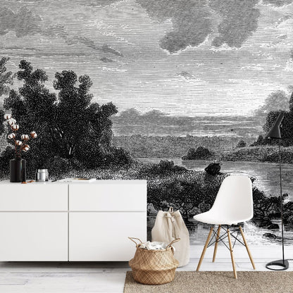 A modern room features a white cabinet with decor items, a white chair, a woven basket, and a black floor lamp. The back wall is adorned with the Mystic Lake Wallpaper Mural by Decor2Go Winnipeg, showcasing a serene nature scene with trees, a river, and a cloudy sky in black and white.