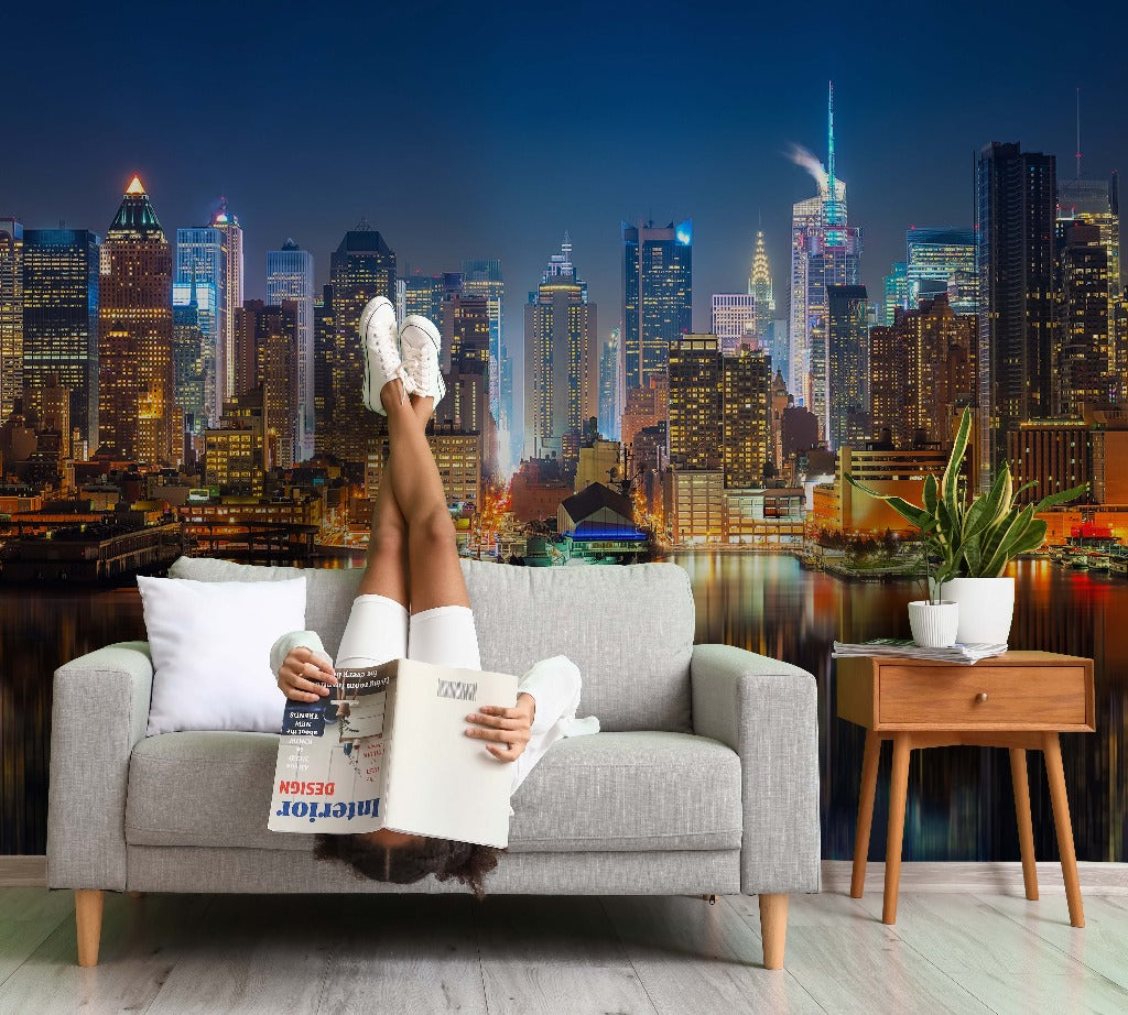 A person reclines on a gray sofa reading "interior design" magazine, feet up in the air against a vibrant Decor2Go Wallpaper Mural at night, with a small side table nearby.