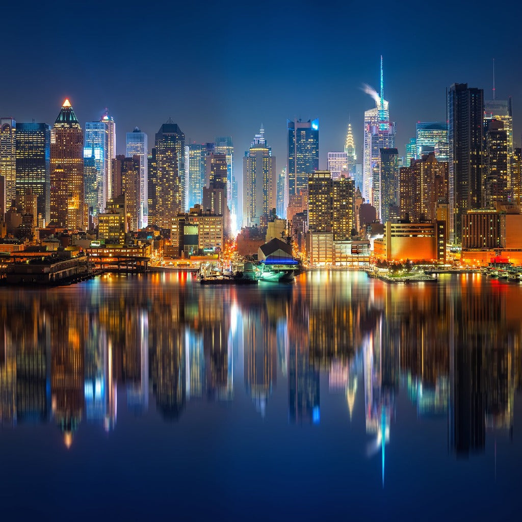 A panoramic night view of the Decor2Go Wallpaper Mural NYC Skyline Wallpaper Mural with illuminated skyscrapers reflecting in the water, showcasing a vibrant urban aesthetic.
