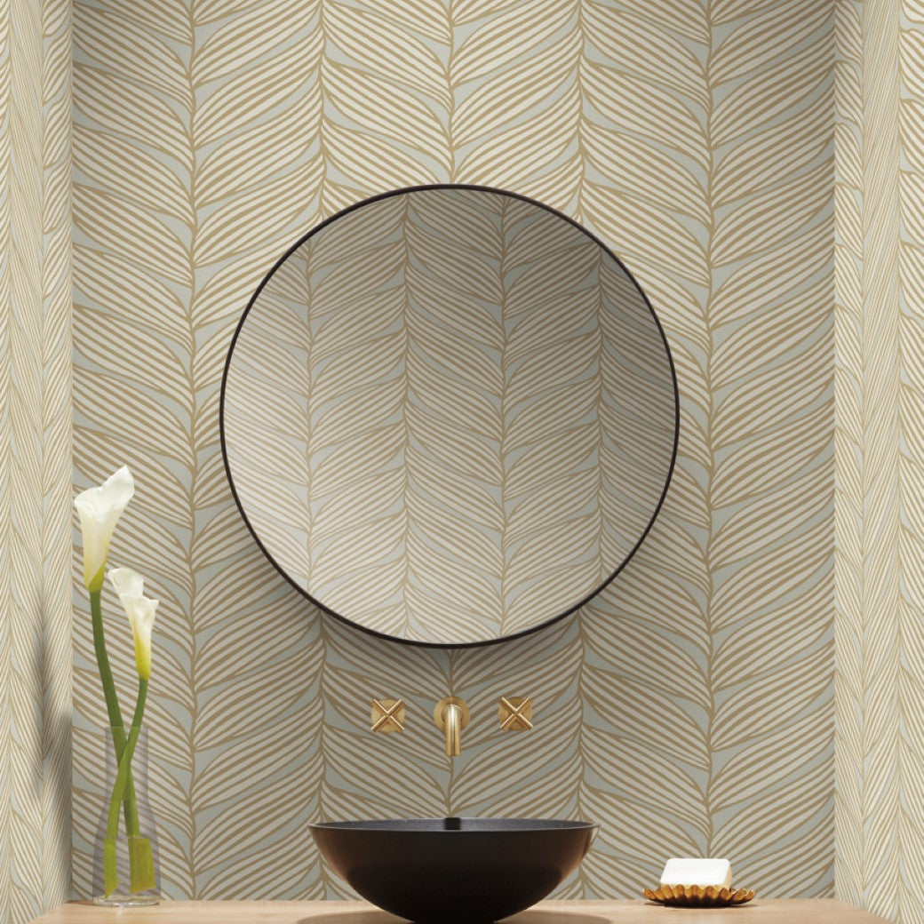 A bathroom has a round mirror above a black vessel sink with a gold faucet from the Modern Metals Collection, set against York Wallcoverings' Neutral & Gold Luminous Leaves Wallpaper. Two white calla lilies in a tall vase to the left add elegance.