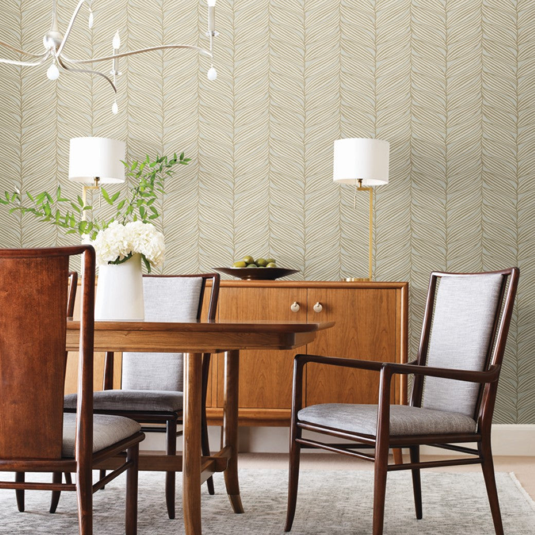 A wooden dining set is enhanced by the Modern Metals Collection, with a metallic finish on the sideboard hosting flowers and lamps. A unique light fixture adds charm, while York Wallcoverings' Neutral & Gold Luminous Leaves Wallpaper subtly accents the room's decor.