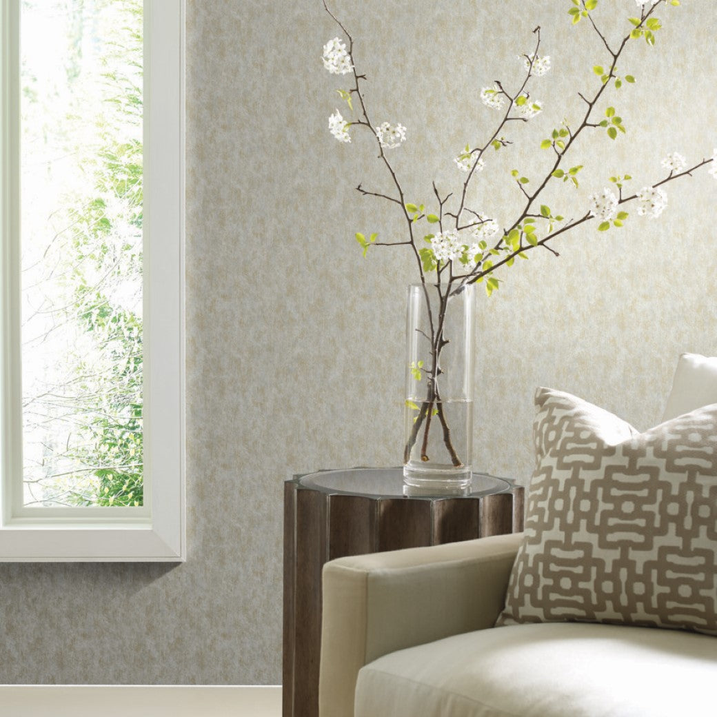 In a modern living room corner, a beige armchair is paired with a geometric decorative pillow. A wooden side table holds a clear vase with blossoming branches, while the Neutral & Gold Shimmering Patina Wallpaper by York Wallcoverings reflects natural light through a large window.