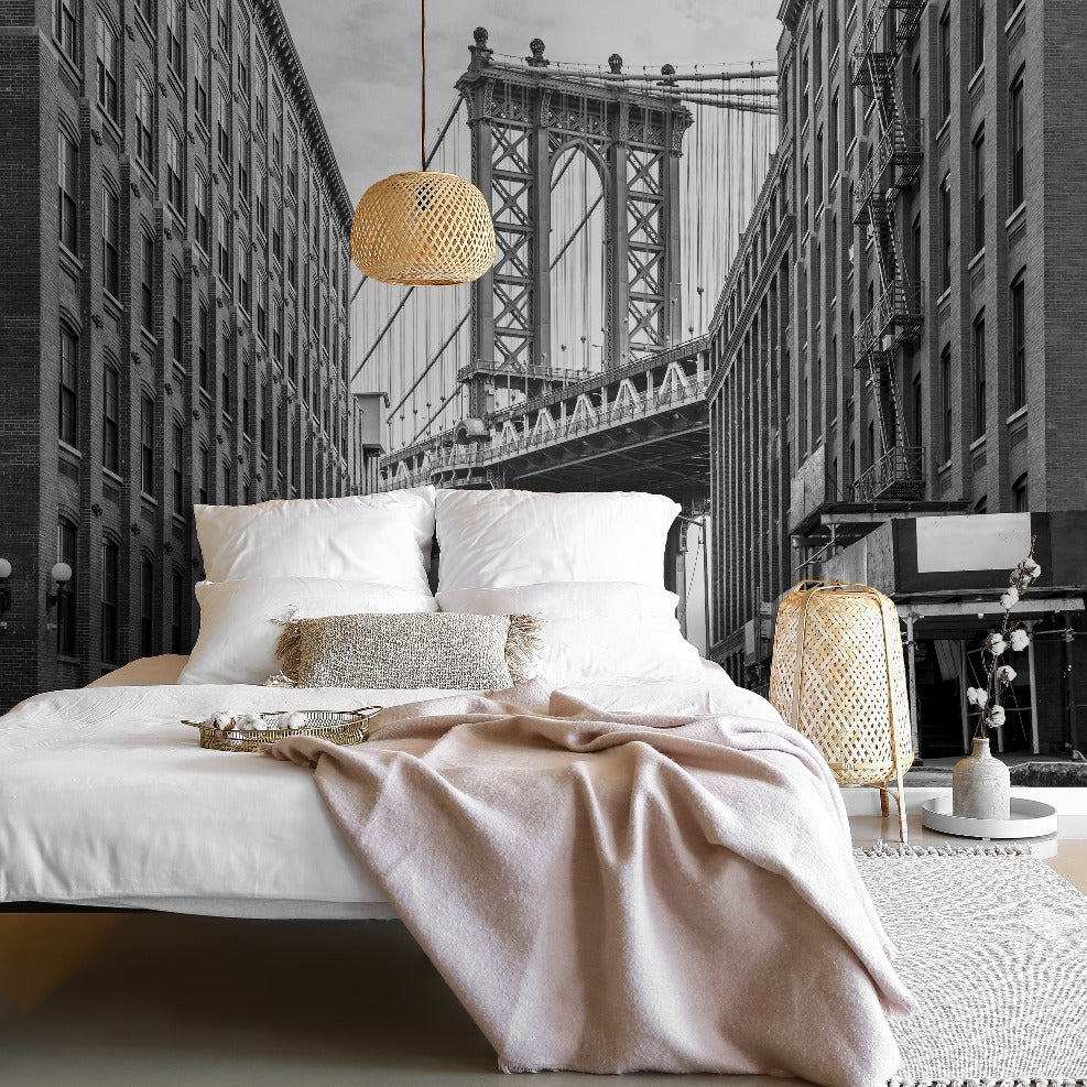A stylish bedroom with a large bed covered in white linens and a pink throw, a New York Bridge Wallpaper Mural - Black and White by Decor2Go Wallpaper Mural as the background, and modern hanging lights.