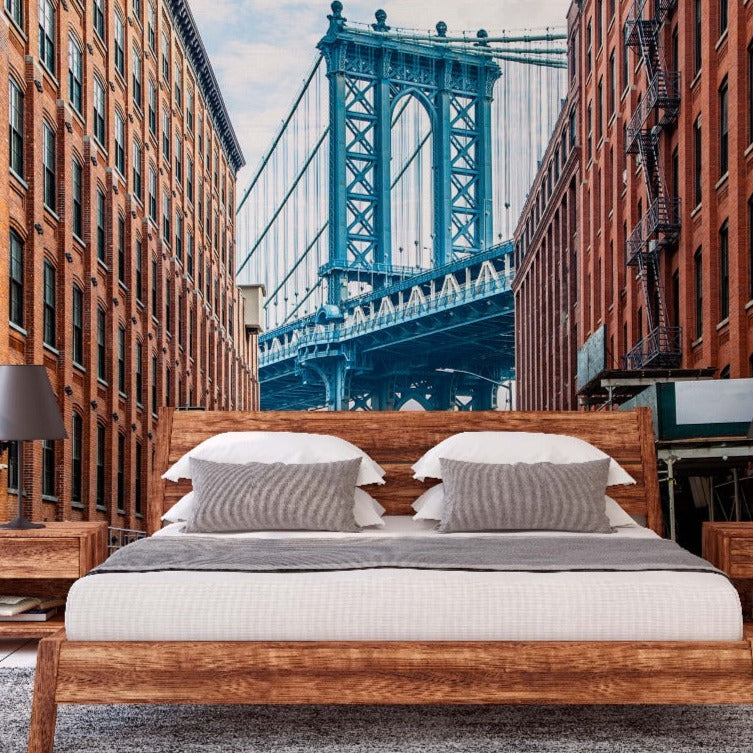 A modern bedroom with a large bed centered between two nightstands, each with a lamp, against a vivid New York Bridge Wallpaper Mural from Decor2Go Wallpaper Mural featuring the Manhattan Bridge and surrounding brick buildings.