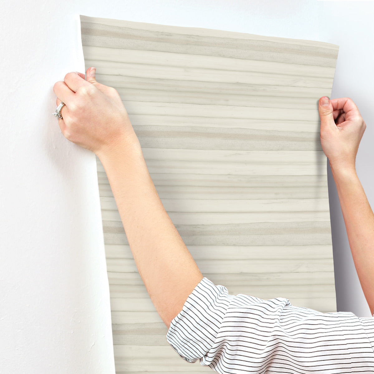 A person wearing a white and black striped shirt is applying York Wallcoverings' Pandora Leaf Oak Wallpaper Brown (60 Sq.Ft.), featuring beige and white stripes, to a wall. They use both hands to smooth and align the wallpaper for easy installation and removal. A silver ring gleams on one of the person's fingers.